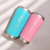 Dos vasos "Termo clásico personalizado 590ml" con nombres personalizados descansan sobre una superficie de madera clara. Un vaso es de color verde azulado y lleva el nombre de Oly Ortiz, y el otro es de color rosa y lleva el nombre de Carolina Ortiz, ambos en elegante letra cursiva. Ambos tienen tapas y bases metálicas que reflejan el encanto clásico de un termo clásico.