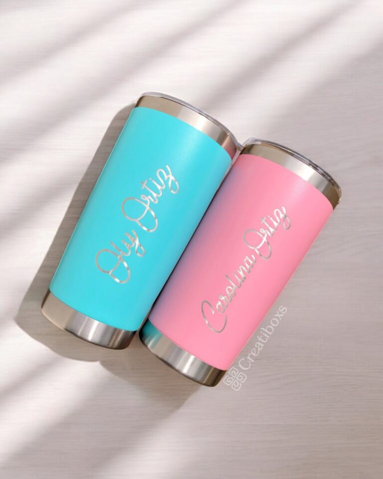 Dos vasos "Termo clásico personalizado 590ml" con nombres personalizados descansan sobre una superficie de madera clara. Un vaso es de color verde azulado y lleva el nombre de Oly Ortiz, y el otro es de color rosa y lleva el nombre de Carolina Ortiz, ambos en elegante letra cursiva. Ambos tienen tapas y bases metálicas que reflejan el encanto clásico de un termo clásico.