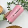 Una persona sostiene un vaso personalizado Termo clásico personalizado de 590 ml y 20 oz en color rosa con texto plateado, rodeado de hojas de eucalipto verde. Se ven parcialmente una estera tejida y un recipiente con tapa de madera. Las palabras Cic.Pahua y Creatiboxs adornan el vaso.