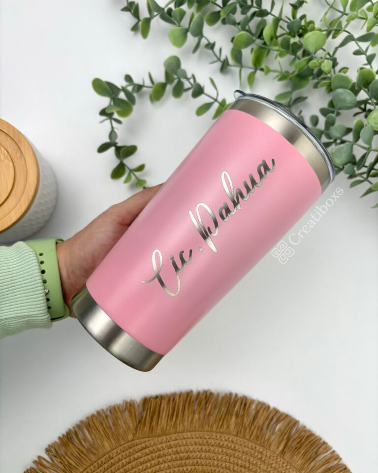 Una persona sostiene un vaso personalizado Termo clásico personalizado de 590 ml y 20 oz en color rosa con texto plateado, rodeado de hojas de eucalipto verde. Se ven parcialmente una estera tejida y un recipiente con tapa de madera. Las palabras Cic.Pahua y Creatiboxs adornan el vaso.
