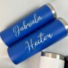 Dos elegantes vasos de metal azules "Termo delgado personalizado 590ml 20oz", personalizados con los nombres "Gabriela" y "Hector" en cursiva, se apilan sobre una superficie blanca rodeada de otros vasos blancos. El nombre de la marca, Creatiboxs, se muestra de forma destacada.