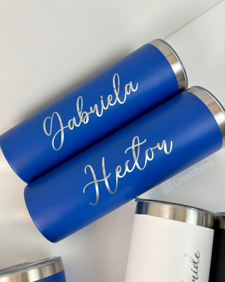 Dos elegantes vasos de metal azules "Termo delgado personalizado 590ml 20oz", personalizados con los nombres "Gabriela" y "Hector" en cursiva, se apilan sobre una superficie blanca rodeada de otros vasos blancos. El nombre de la marca, Creatiboxs, se muestra de forma destacada.