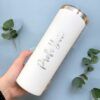 Una mano sostiene un elegante Termo Delgado Personalizado de 590 ml y 20 oz con letras metálicas que dicen Maestra Yuri. Este vaso tiene una tapa y, en el fondo, dos ramitas de vegetación descansan sobre una superficie azul claro.