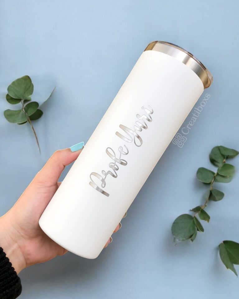 Una mano sostiene un elegante Termo Delgado Personalizado de 590 ml y 20 oz con letras metálicas que dicen Maestra Yuri. Este vaso tiene una tapa y, en el fondo, dos ramitas de vegetación descansan sobre una superficie azul claro.