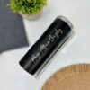 Un Termo Delgado personalizado de 590 ml y 20 oz en color negro, con la inscripción "Hugo Alfonso González" elegantemente escrita en cursiva blanca, reposa sobre una superficie blanca. Acompañando la escena hay una planta en maceta verde, un cuaderno negro cerrado y un mantel circular tejido que aportan una sutil elegancia al arreglo.