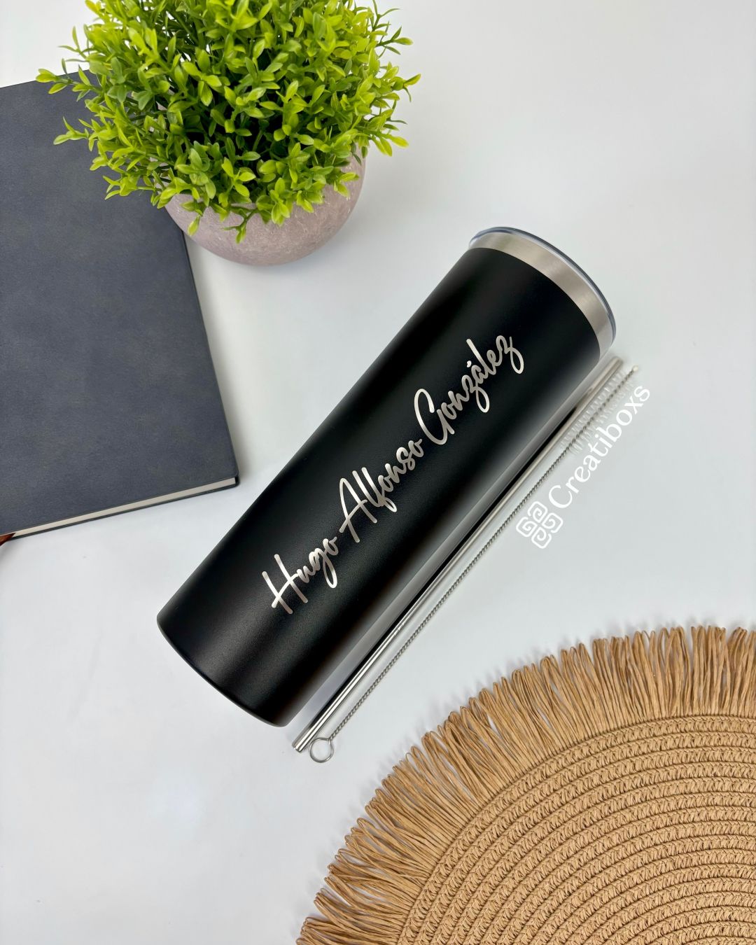 Un Termo Delgado personalizado de 590 ml y 20 oz en color negro, con la inscripción "Hugo Alfonso González" elegantemente escrita en cursiva blanca, reposa sobre una superficie blanca. Acompañando la escena hay una planta en maceta verde, un cuaderno negro cerrado y un mantel circular tejido que aportan una sutil elegancia al arreglo.