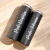 Sobre una superficie de madera, dos vasos Termo Delgado personalizado de 590 ml con bordes plateados se encuentran elegantemente colocados. Estos vasos térmicos negros presentan un texto personalizado en cursiva blanca que muestra los nombres Profe Drazitza y Profe Adriana, lo que resalta su elegante diseño.