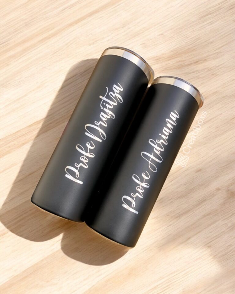 Sobre una superficie de madera, dos vasos Termo Delgado personalizado de 590 ml con bordes plateados se encuentran elegantemente colocados. Estos vasos térmicos negros presentan un texto personalizado en cursiva blanca que muestra los nombres Profe Drazitza y Profe Adriana, lo que resalta su elegante diseño.