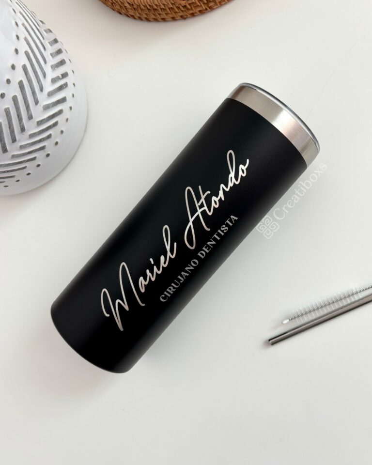 Un termo delgado personalizado negro de 590 ml y 20 oz muestra "Mariel Atando Cirujano Dentista" en una elegante letra cursiva blanca. Cerca de la superficie blanca, encontrará una pajita de metal y un objeto blanco texturizado.