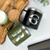 Sobre una superficie de madera adornada con hojas verdes, hay dos tazas. Una es una "Termo taza mediana" personalizada de color negro con la letra G y la palabra GIGIS inscritas. La otra es una taza verde de 420 ml con la letra R y la palabra BILLIE. Cerca, también hay un posavasos de corcho y una vela en un portavelas verde.