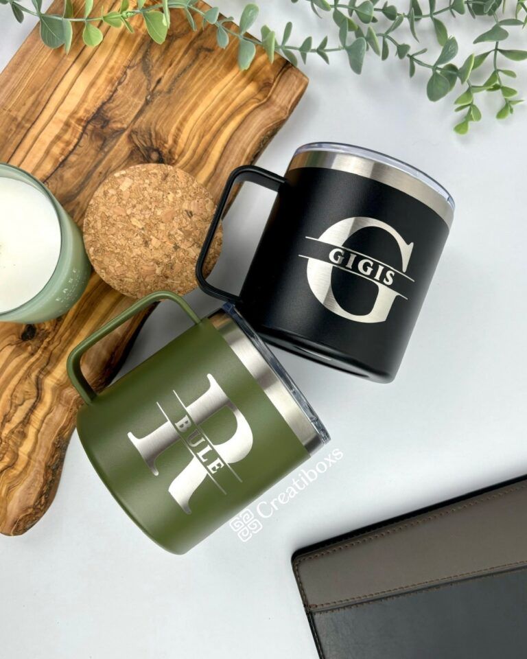 Sobre una superficie de madera adornada con hojas verdes, hay dos tazas. Una es una "Termo taza mediana" personalizada de color negro con la letra G y la palabra GIGIS inscritas. La otra es una taza verde de 420 ml con la letra R y la palabra BILLIE. Cerca, también hay un posavasos de corcho y una vela en un portavelas verde.
