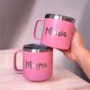 Se muestran dos tazas personalizadas Termo Taza Mediano de color rosa con tapas plateadas. Una taza, sostenida en una mano, tiene el nombre María, mientras que la otra elegante taza reposa sobre una superficie de madera y está adornada con el nombre Mayra.