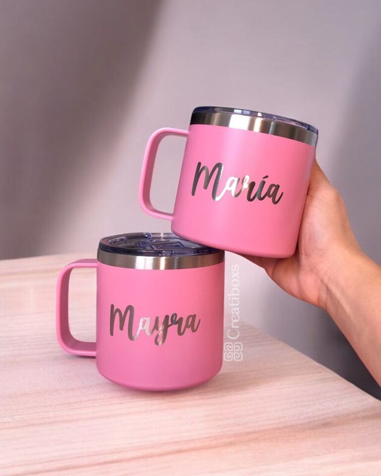Se muestran dos tazas personalizadas Termo Taza Mediano de color rosa con tapas plateadas. Una taza, sostenida en una mano, tiene el nombre María, mientras que la otra elegante taza reposa sobre una superficie de madera y está adornada con el nombre Mayra.