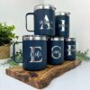 Se muestran cinco tazas personalizadas Termo taza de 360 ml de color azul marino con tapas plateadas sobre una tabla de madera. Cada taza personalizada muestra una inicial grande y un nombre: Arnulfo, Isabel, Erika, Gabby y Fernando. Estas elegantes tazas añaden encanto a cualquier entorno con plantas verdes de fondo.