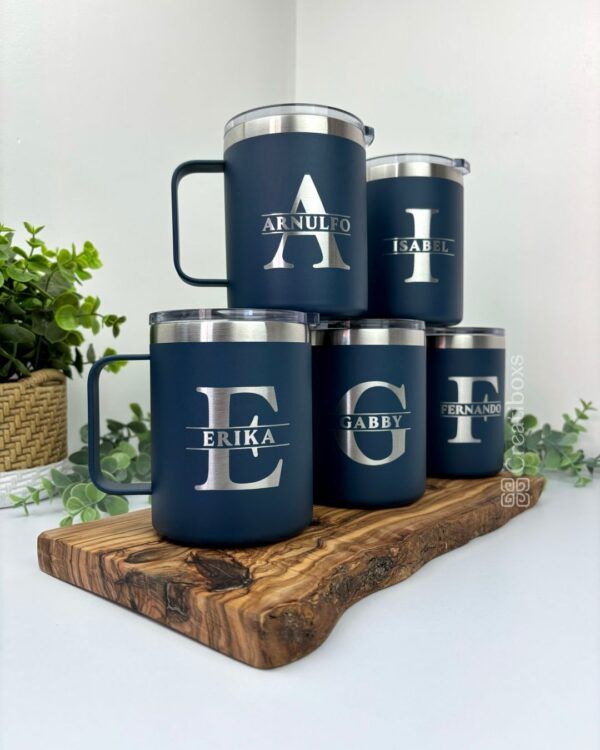 Se muestran cinco tazas personalizadas Termo taza de 360 ml de color azul marino con tapas plateadas sobre una tabla de madera. Cada taza personalizada muestra una inicial grande y un nombre: Arnulfo, Isabel, Erika, Gabby y Fernando. Estas elegantes tazas añaden encanto a cualquier entorno con plantas verdes de fondo.