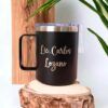 Termo taza personalizada de 360 ml 12 oz con la inscripción Lic. Carlos Lozano, sobre una superficie de madera, con tapa transparente y asa negra. Esta taza de metal negra personalizada está rodeada de hojas verdes sobre un fondo de paneles de madera verticales.