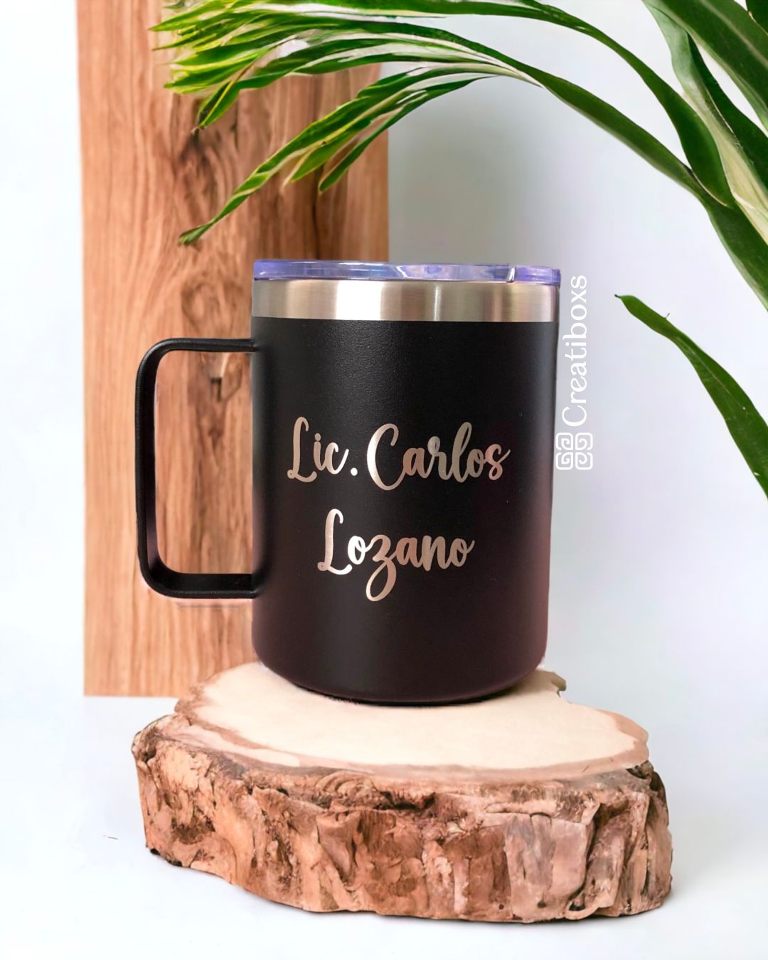 Termo taza personalizada de 360 ml 12 oz con la inscripción Lic. Carlos Lozano, sobre una superficie de madera, con tapa transparente y asa negra. Esta taza de metal negra personalizada está rodeada de hojas verdes sobre un fondo de paneles de madera verticales.