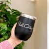 Una mano sostiene un elegante termo vinero personalizado de 360 ml y 12 oz de color negro con la inscripción "Pail Ortega" en él. El vaso tiene un borde plateado y está decorado con un fondo de hojas verdes exuberantes y una pared blanca prístina.