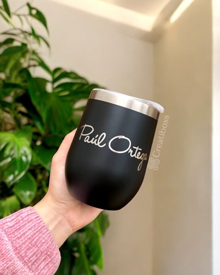 Una mano sostiene un elegante termo vinero personalizado de 360 ml y 12 oz de color negro con la inscripción "Pail Ortega" en él. El vaso tiene un borde plateado y está decorado con un fondo de hojas verdes exuberantes y una pared blanca prístina.