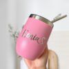 Una mano sostiene un Termo Vinero Personalizado rosa con una capacidad de 360 ml o 12 oz, con borde metálico y pajita. El nombre Kenia, acompañado por un símbolo de corazón, está bellamente inscrito en letras plateadas en el vaso. Un fondo de cocina difuminado brinda un ambiente acogedor.