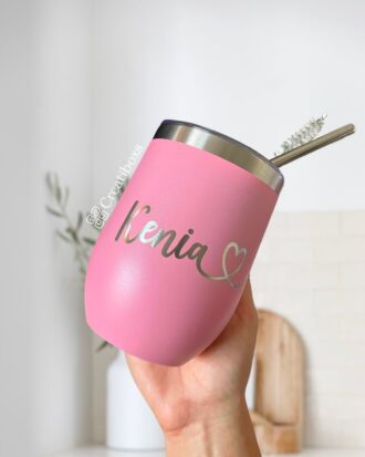 Una mano sostiene un Termo Vinero Personalizado rosa con una capacidad de 360 ml o 12 oz, con borde metálico y pajita. El nombre Kenia, acompañado por un símbolo de corazón, está bellamente inscrito en letras plateadas en el vaso. Un fondo de cocina difuminado brinda un ambiente acogedor.