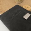 Libreta personalizada A5 con broche magnético plateado, negra, con el nombre José Carlos Diaz, Gerente de Recursos Humanos, grabado. Sobre un tapete tejido junto a una planta.