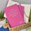 Libreta personalizada A5 lisa con elástico en color rosa y frase en español, colocada en una cesta de mimbre junto a plantas y diarios abiertos.