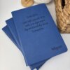 Tres Libreta personalizada azul A5 lisa con elástico apiladas sobre una superficie blanca, cada una con "Miguel" y una cita motivadora en la portada.