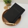 A Libreta personalizada A5 lisa con elástico en color negro descansa en una canasta de mimbre, junto a una pequeña maceta sobre una superficie blanca.