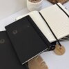 Tres cuadernos negros, entre ellos una Libreta personalizada A5 lisa con elástico -uno abierto con páginas en blanco- están colocados junto a una suculenta maceta sobre una alfombra tejida.