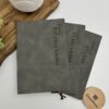 Tres libretas "Libreta personalizada A5 lisa tacto suave" de color gris, con nombres impresos en vertical, se exponen sobre una superficie blanca junto a una bandeja de madera.