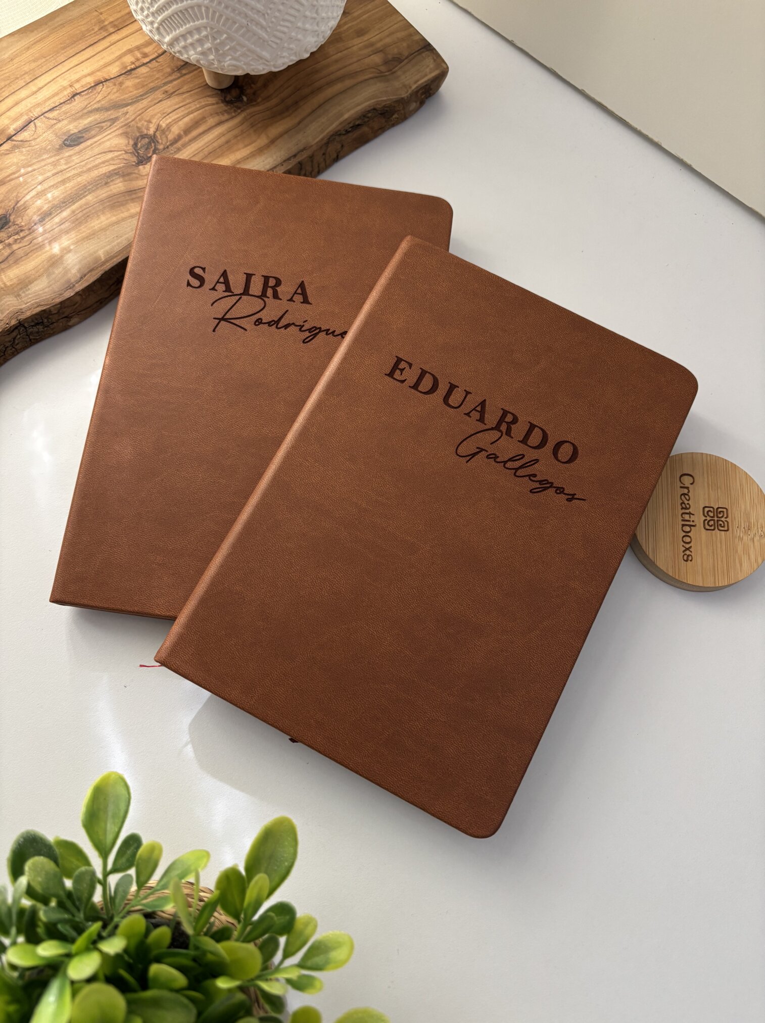 Dos "Libreta personalizada A5 lisa con elástico" de color marrón, personalizadas con los nombres de Saira Rodríguez y Eduardo Gallegos, descansan sobre un escritorio blanco decorado con plantas.