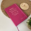 Una Libreta personalizada A5 lisa con elástico rosa y texto en español descansa sobre un tapete tejido, junto a un portavasos de madera y una pequeña planta verde.
