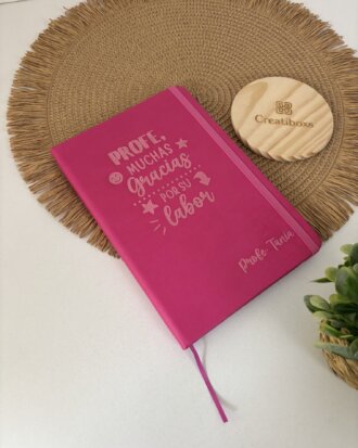 Una Libreta personalizada A5 lisa con elástico rosa y texto en español descansa sobre un tapete tejido, junto a un portavasos de madera y una pequeña planta verde.