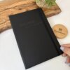 Una libreta personalizada A5 lisa con elástico y el nombre ING. MEP Luis Rojas en la portada, acompañada de un posavasos de madera Creatiboxs y una planta.
