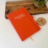 A Libreta personalizada A5 lisa con elástico con la etiqueta OLIVER Restaurant descansa sobre una superficie blanca junto a una planta y una bandeja de madera.