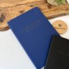 Libreta personalizada A5 lisa con elástico azul y texto dorado sobre una mesa, junto a una libreta negra, una planta y un posavasos de madera Creatiboxs.