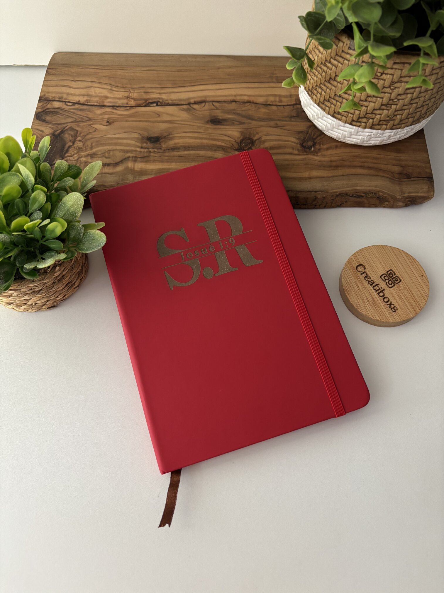 Una libreta personalizada A5 lisa con elástico en color rojo y "S.R. Josué" en la portada, rodeada de plantas en macetas sobre una superficie blanca.