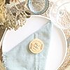 Un plato blanco redondo con una servilleta texturizada de color azul claro, un abridor de botellas de bambú redondo personalizado del "Paquete de 10 - 50 Destapador con imán redondo de bambú personalizado", además de un tazón transparente, una copa de vino y flores secas sobre un mantel individual tejido.