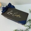 Kit termo taza 710ml y caja chica personalizada en color negro con “Leo Castro” grabado en dorado, decorado con listón azul marino y acompañado de hojas verdes.