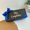 Una caja de regalo negra con una cinta azul, que presenta el Kit termo taza 710ml y caja chica personalizada, dice "Feliz Cumpleaños" sobre un fondo blanco junto a un mantel individual.