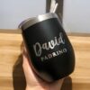 Una mano sostiene un elegante vaso Duo Padrinos negro con borde plateado que muestra el nombre David Padrino en nítidas letras blancas. Una pajita atraviesa la tapa de este termo vinero personalizado de 360 ml. La luz del sol baila sobre la superficie de madera y proyecta sombras divertidas en el fondo.