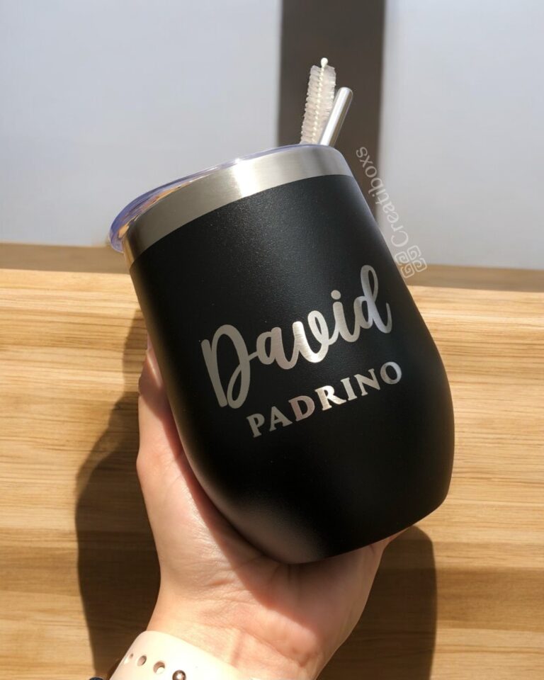 Una mano sostiene un elegante vaso Duo Padrinos negro con borde plateado que muestra el nombre David Padrino en nítidas letras blancas. Una pajita atraviesa la tapa de este termo vinero personalizado de 360 ml. La luz del sol baila sobre la superficie de madera y proyecta sombras divertidas en el fondo.