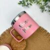 Una taza rosa con la palabra "Alessa Mona" personalizada mediante grabado láser reposa sobre un mantel individual tejido. Cerca, un pequeño recipiente rosa con tapa de corcho complementa la escena, mientras que la parte superior de la imagen está decorada con vegetación.
