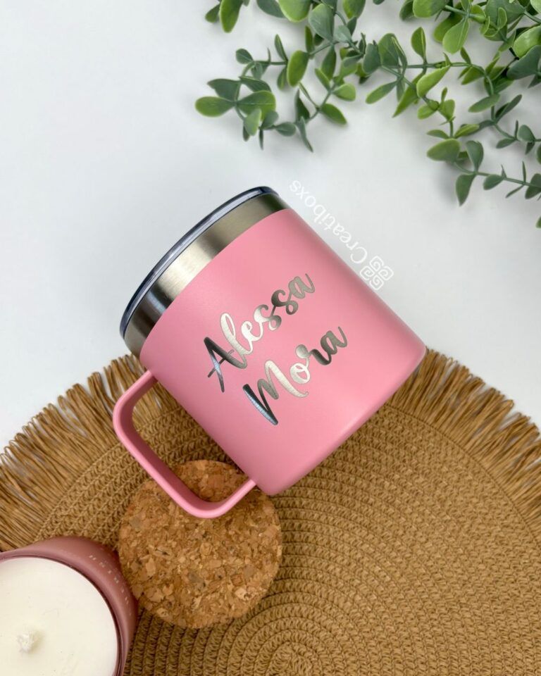 Una taza rosa con la palabra "Alessa Mona" personalizada mediante grabado láser reposa sobre un mantel individual tejido. Cerca, un pequeño recipiente rosa con tapa de corcho complementa la escena, mientras que la parte superior de la imagen está decorada con vegetación.