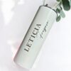 Un deslumbrante vaso cilíndrico metálico, etiquetado como "Termo personalizado glitter 590ml 20oz", reposa de lado con el nombre LETICIA Fregoso en letras negras en negrita. Un cepillo de limpieza y una ramita de eucalipto emergen de su abertura, mientras que un fondo suavemente iluminado proyecta sombras suaves, amplificando su encanto.