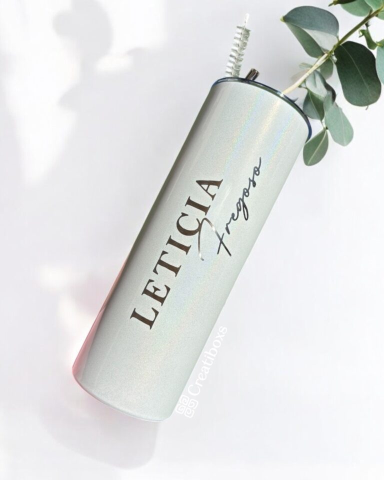Un deslumbrante vaso cilíndrico metálico, etiquetado como "Termo personalizado glitter 590ml 20oz", reposa de lado con el nombre LETICIA Fregoso en letras negras en negrita. Un cepillo de limpieza y una ramita de eucalipto emergen de su abertura, mientras que un fondo suavemente iluminado proyecta sombras suaves, amplificando su encanto.