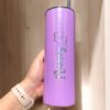 Un Termo Personalizado Glitter de 590 ml en color lavanda, sostenido en posición vertical por una mano que lleva un reloj inteligente beige, muestra el nombre Nancy Pérez en cursiva, adornado con un gráfico de corazón. Dentro del vaso hay una pajita de acero inoxidable.