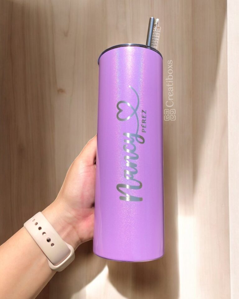 Un Termo Personalizado Glitter de 590 ml en color lavanda, sostenido en posición vertical por una mano que lleva un reloj inteligente beige, muestra el nombre Nancy Pérez en cursiva, adornado con un gráfico de corazón. Dentro del vaso hay una pajita de acero inoxidable.