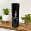 Un elegante termo personalizado liso de color negro de 590 ml y 20 oz con el nombre Iván grabado sobre una superficie de madera. Este vaso está rodeado de pequeñas plantas y cuenta con una pajita de metal, lo que crea una escena armoniosa contra el fondo blanco limpio.